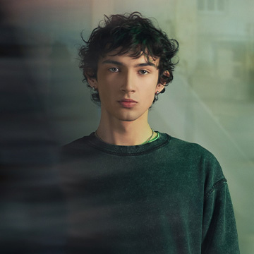 A person standing indoors near a window, wearing a sweater. The background shows a blurred outside view, suggesting a calm and reflective atmosphere.