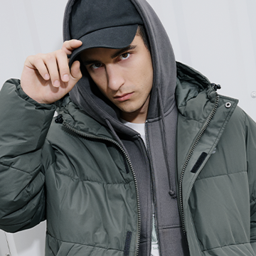 A person wearing a dark gray hoodie underneath a thick padded jacket with a high collar and front zipper. One hand is adjusting the hood of the jacket. The outfit appears suitable for cold weather. banner