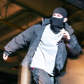 A person dressed in outdoor winter clothing, including a jacket and a balaclava, appears to be running in an urban environment, possibly under a structure or building with industrial elements. banner