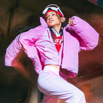 A person is captured mid-dance in energetic motion, wearing a puffy jacket and sporty attire. They appear to be outdoors, exuding a sense of dynamism and liveliness. banner