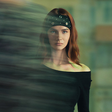 A person is standing indoors, wearing an off-the-shoulder black top. A blurred figure passes in front, partially obstructing the view. The image captures a moment of movement and stillness in a casual environment.