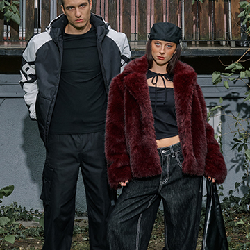 Two people are standing outdoors, dressed in trendy, casual outfits. One is wearing a black jacket and pants, while the other is in a burgundy faux fur jacket, black top, jeans, and holding a black handbag. They are surrounded by greenery and a wooden fence backdrop. banner