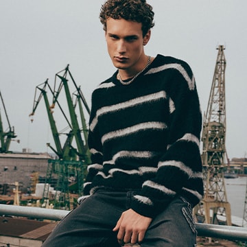 Man wearing a striped sweater and dark trousers in an industrial port setting, presenting a casual urban fashion style